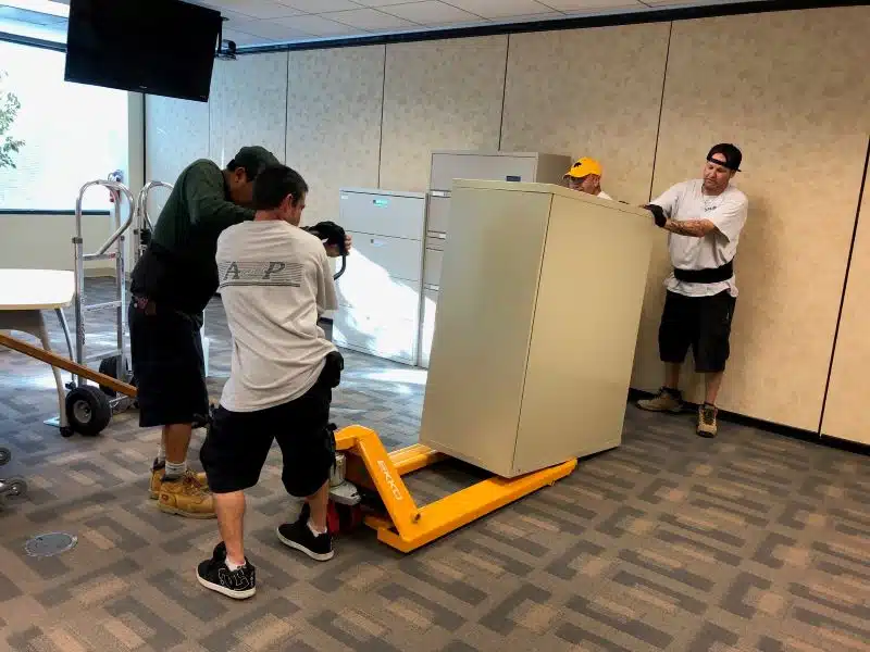 A team of A and P employees are loading a large fireproof file cabinet onto a hydraulic dolly.