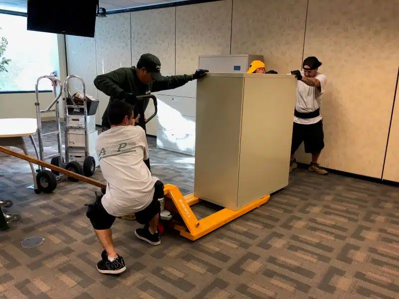 A team of A and P employees are loading a large fireproof file cabinet onto a hydraulic dolly.