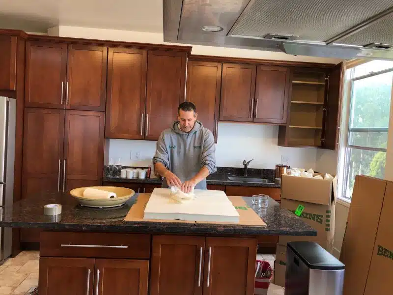 A person wearing an A and P branded sweatshirt is carefully packing up glass items in a kitchen using tissue paper.