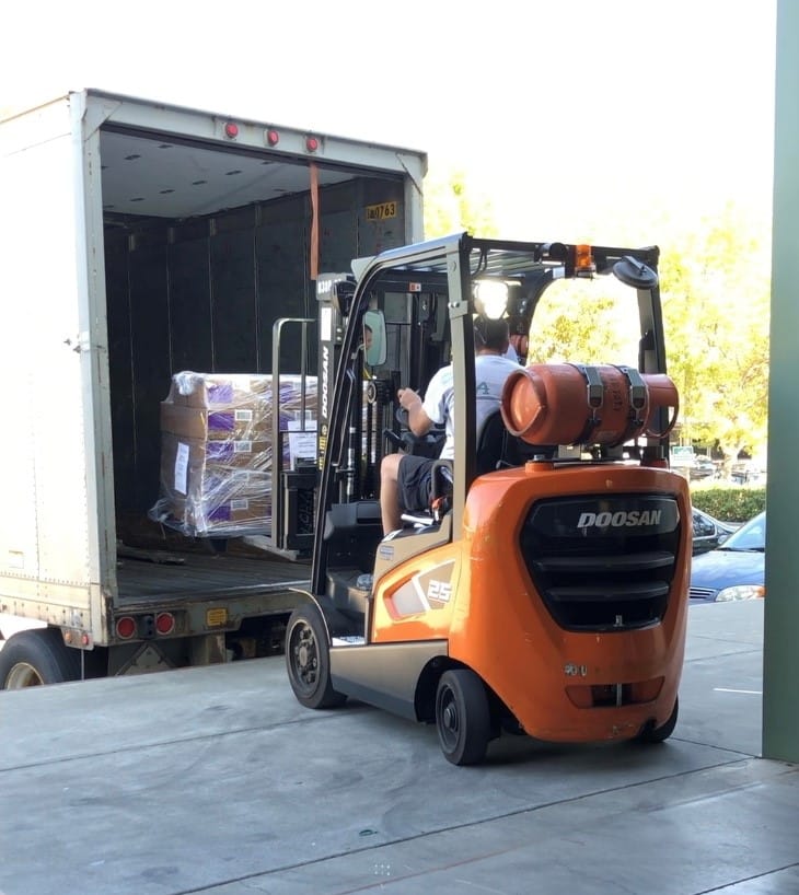 A driver uses a forklift to carefully load a wrapped pallet into a truck at a loading dock.