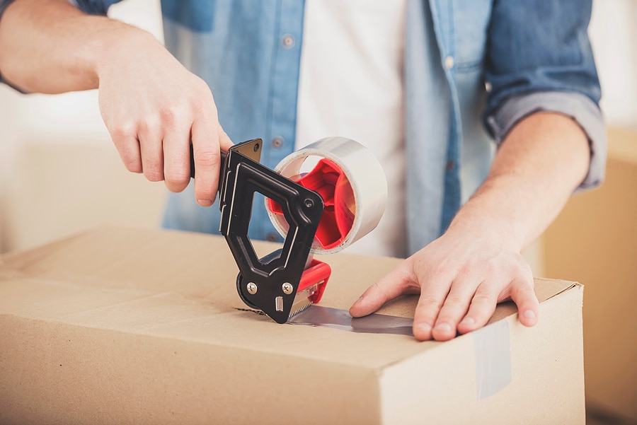 A person is sealing up a cardboard moving box by applying a strip of packing tape across the top center.